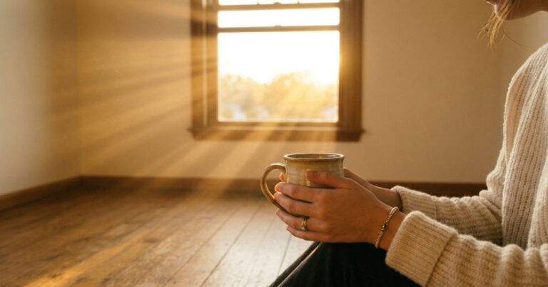 A blonde woman seen from behind holding a coffee mug at a sunlit desk with an open notebook beside her.