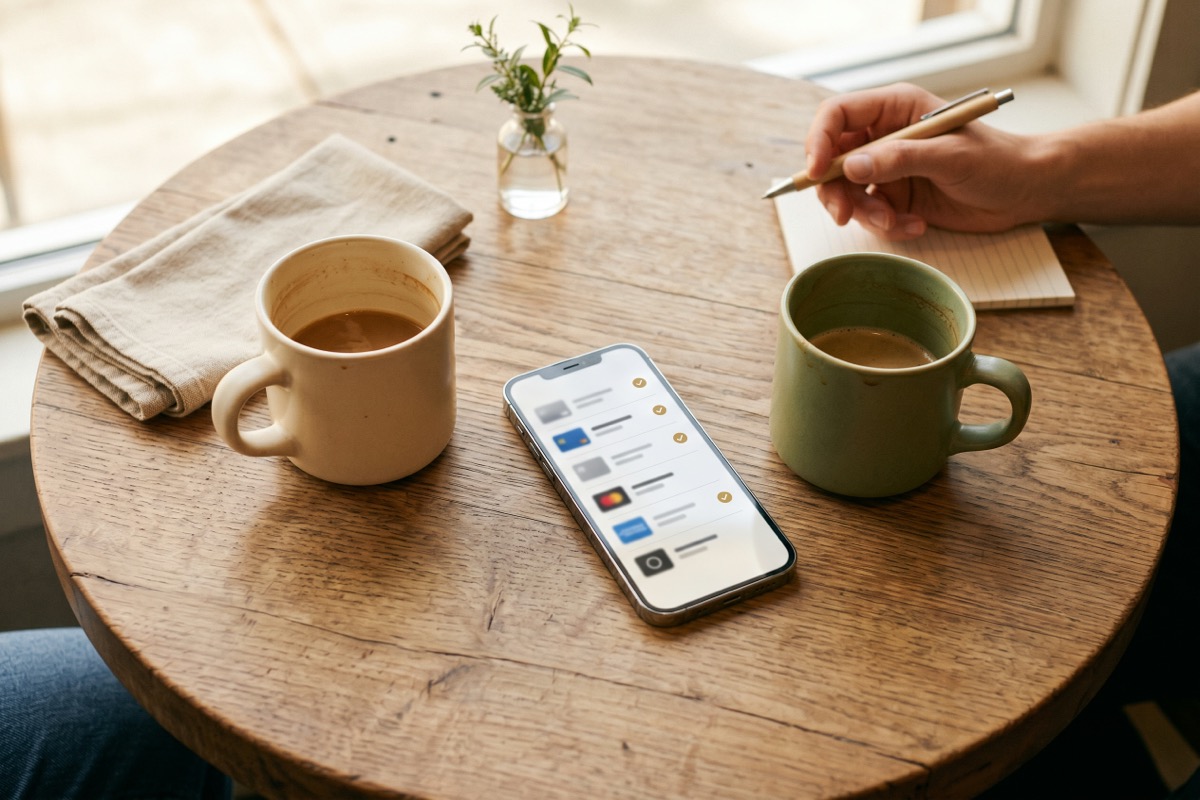 Top-down view of two coffee mugs on a wooden cafe table with a phone between them showing a list of subscription perks marked with golden checkmarks.