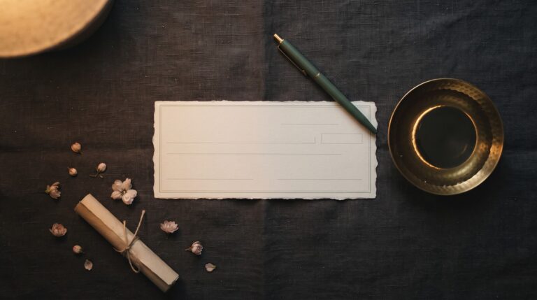 A blank cream paper check and a sage-barreled pen on dark charcoal linen beside a small brass dish of still dark water rimmed in gold like a new moon.