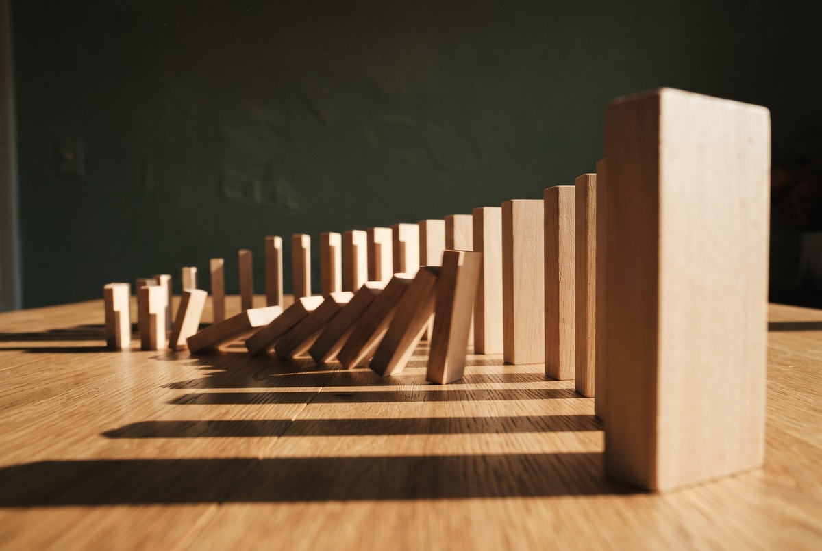 A line of plain wooden blocks standing like dominoes on a table, growing from small to large, with the small ones at the start tipping over toward the bigger ones